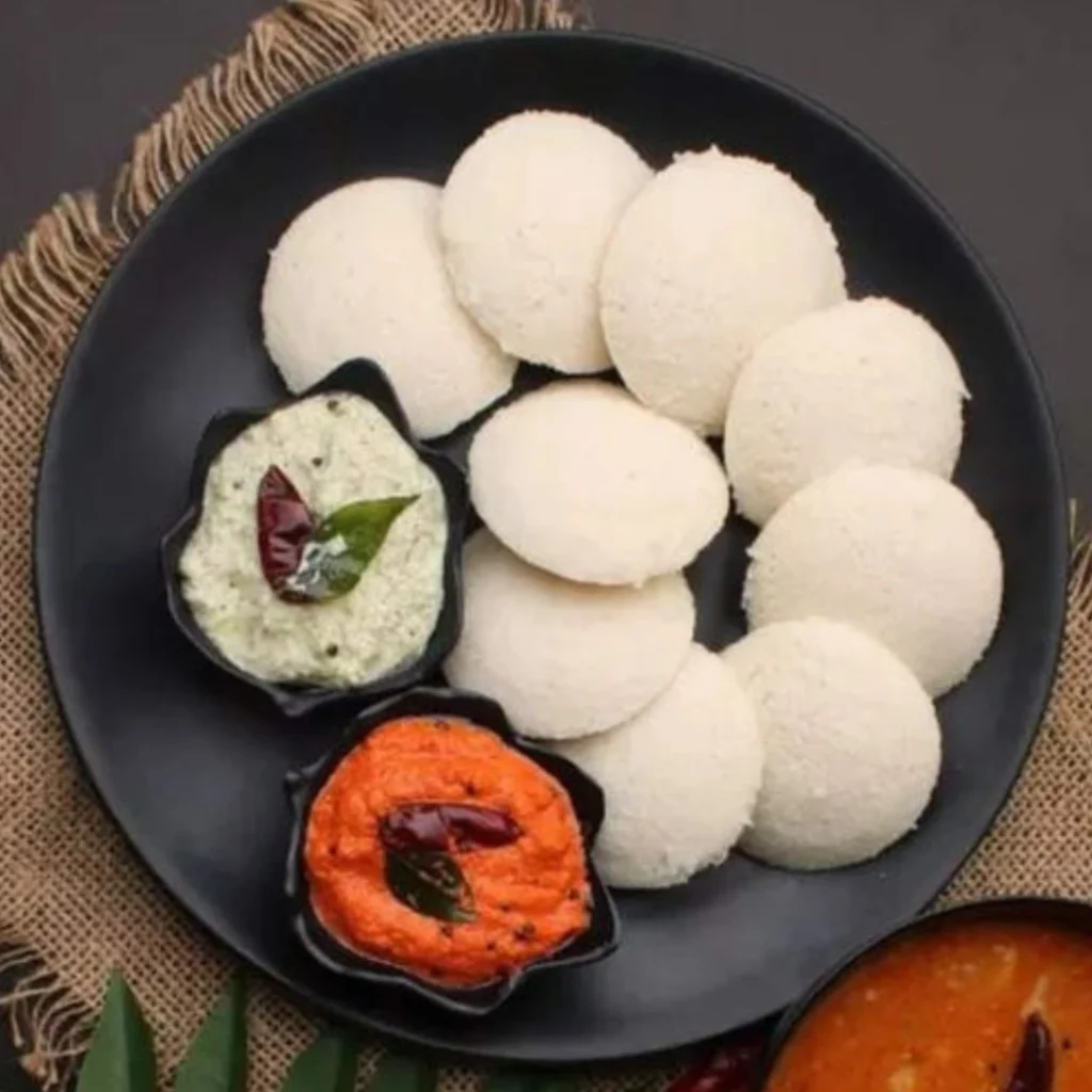 South Indian idli with coconut chutney and red tomato chutney served on black plate with curry leaves garnish