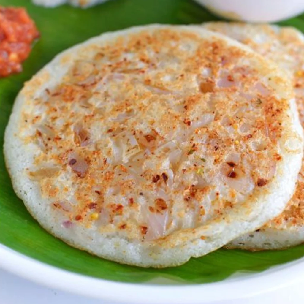 "Onion uthappam served on banana leaf with spicy red chutney, traditional South Indian rice and lentil pancake with chopped onions and spices"