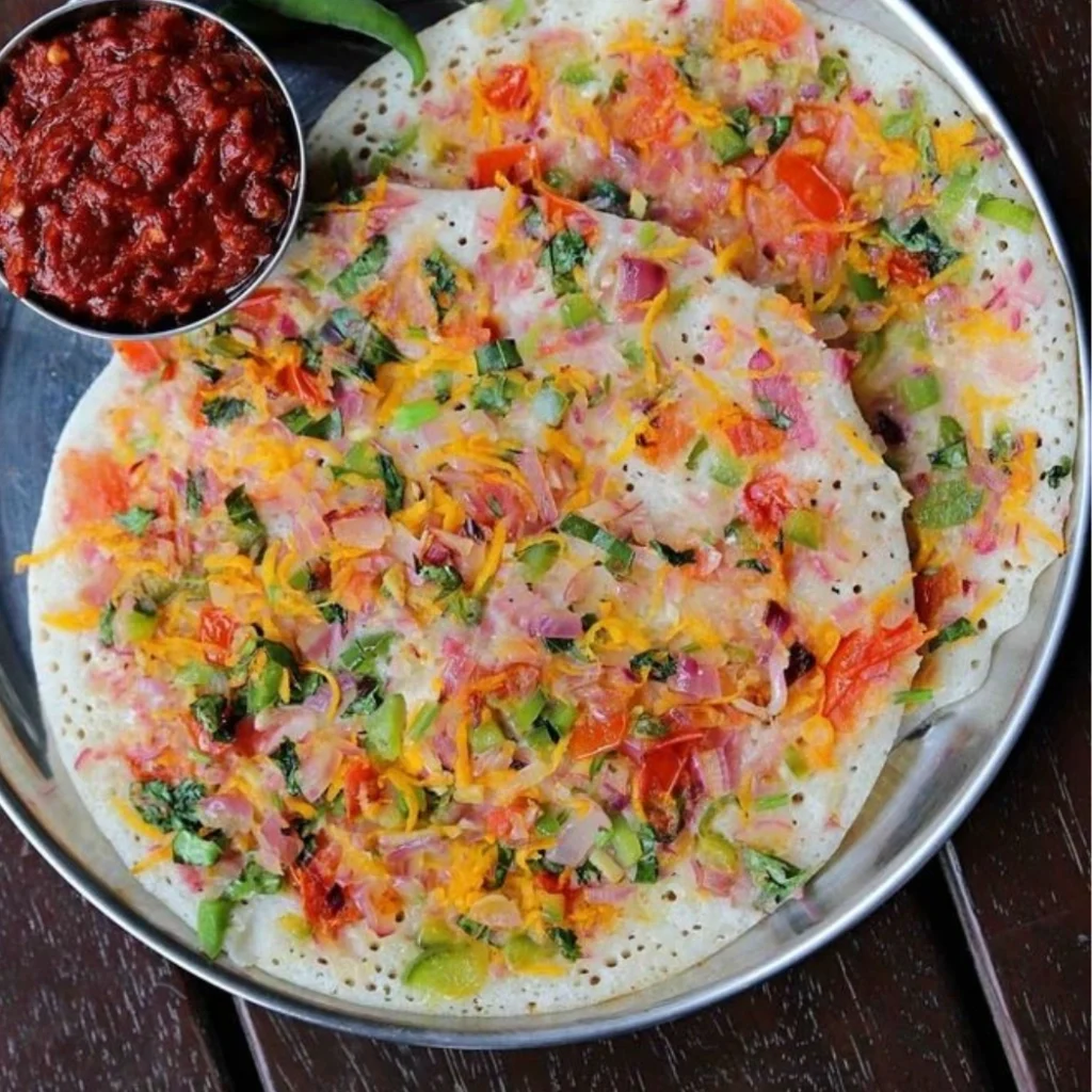 "Homemade Rava dosa topped with colorful vegetables including onions, tomatoes, carrots, and green chilies, served with red chutney in a steel plate"