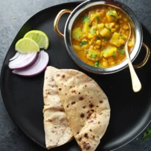 "Aloo curry served with roti, sliced red onions and lime wedges on a black plate - traditional Indian vegetarian meal"