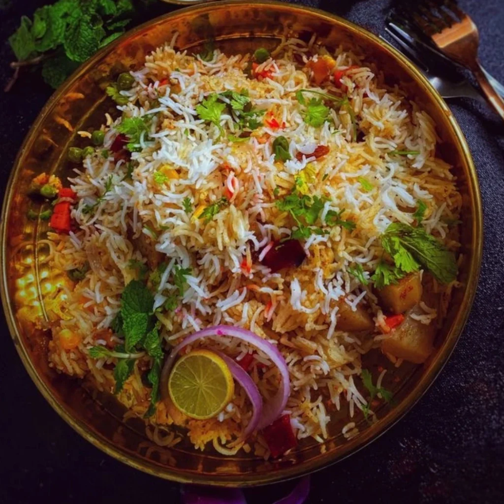 "Aromatic Indian vegetable biryani served in brass bowl garnished with fresh herbs, lime and red onions"