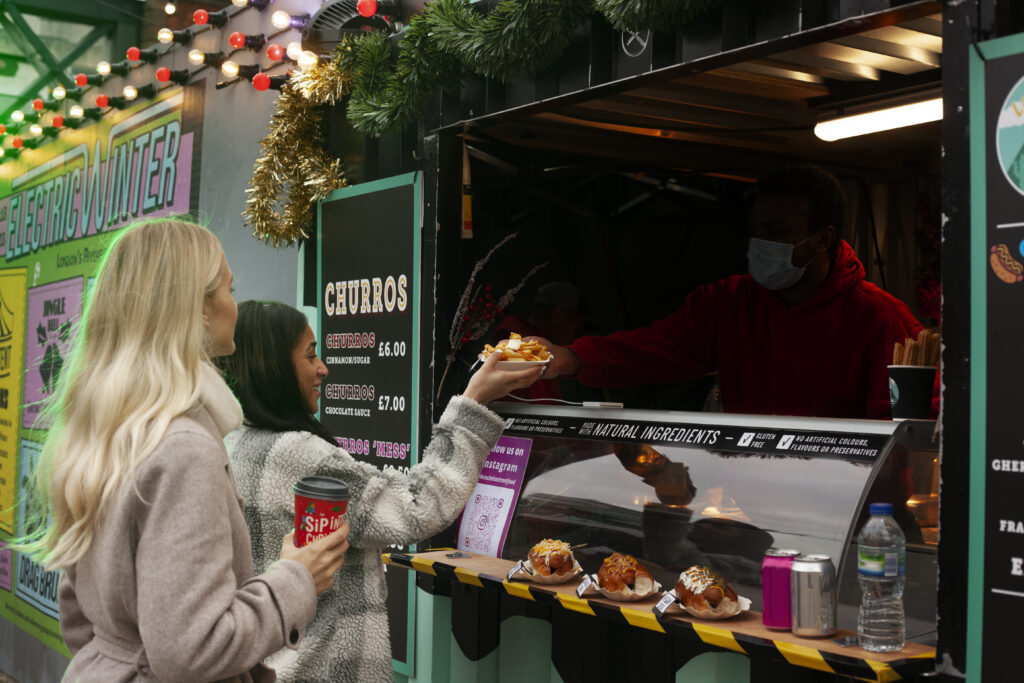 "Two women buying churros from a festive food stall in London with Christmas decorations and lights"